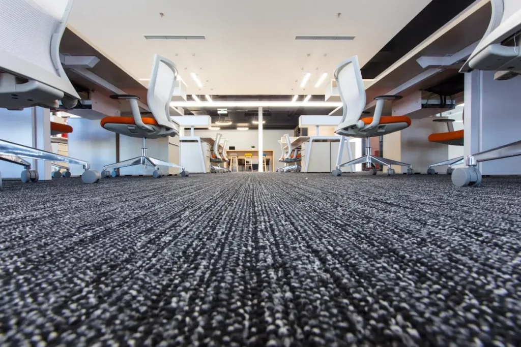 A modern office space featuring rows of white rolling chairs with orange cushions at long desks. The floor is covered with a textured gray carpet, and the ceiling above displays bright lights.