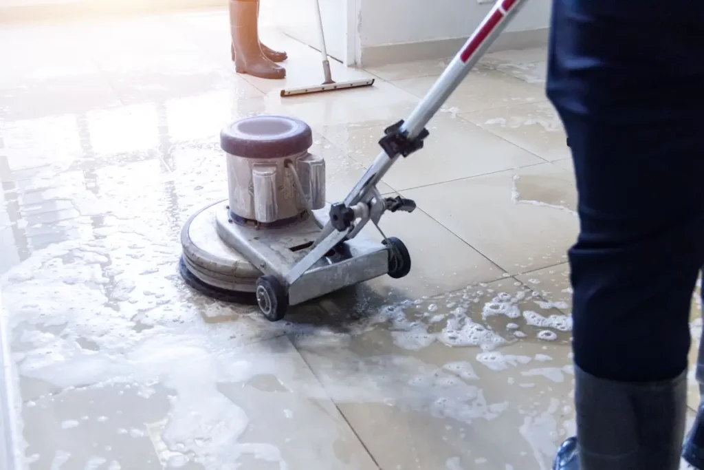 A person cleaning a tiled floor with a floor scrubber, creating soapy suds. Another person in the background uses a squeegee to remove water. The scene suggests a commercial or industrial cleaning process.