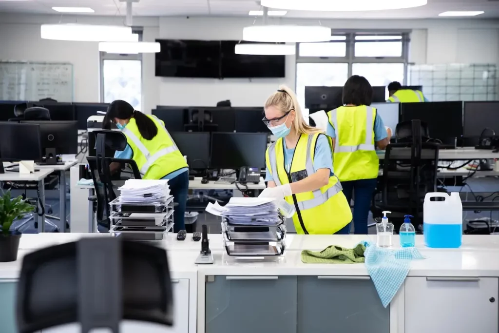 Workers wearing masks and yellow safety vests clean an office. They sanitize desks and organize papers. Cleaning supplies are on the desk in the foreground. Computers and chairs are visible throughout the office.