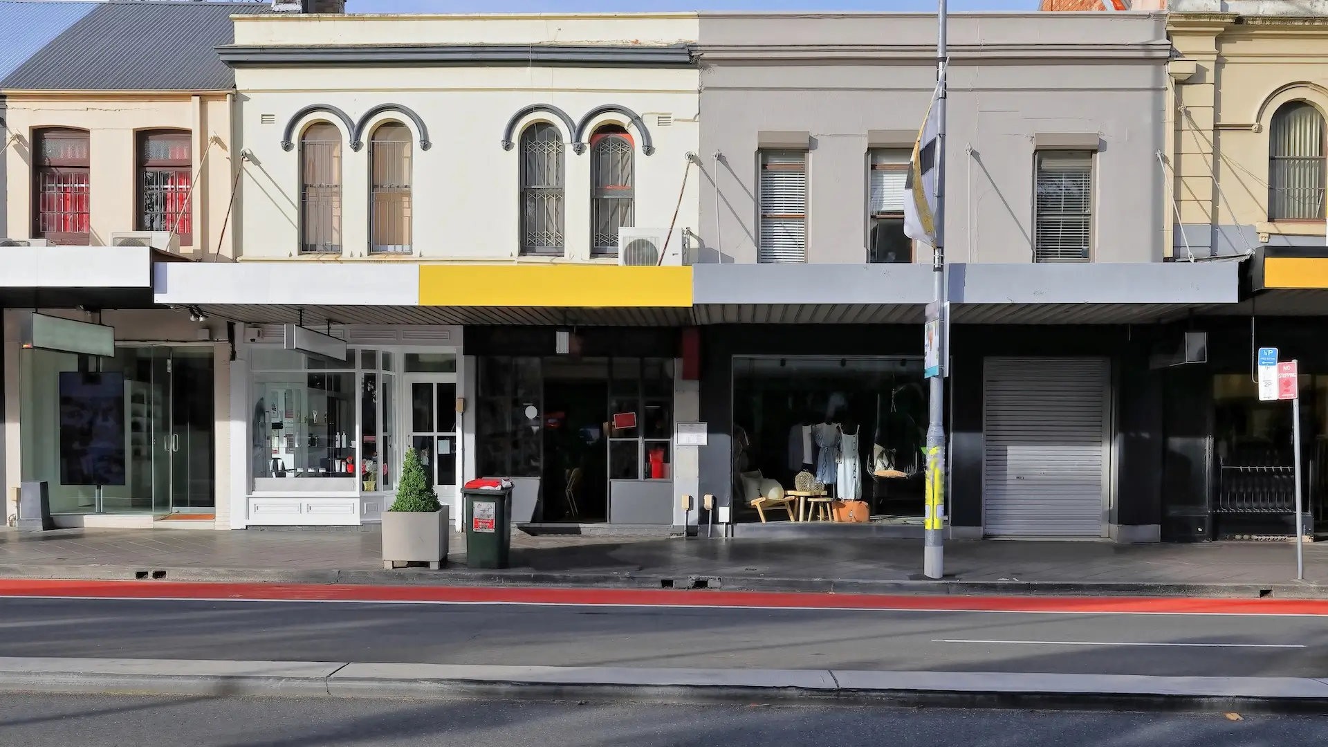 Street view of a row of buildings with shops. The facades have large windows and awnings. There's a red line on the pavement, and a gray building with a closed shutter on the right. A pole stands on the sidewalk.