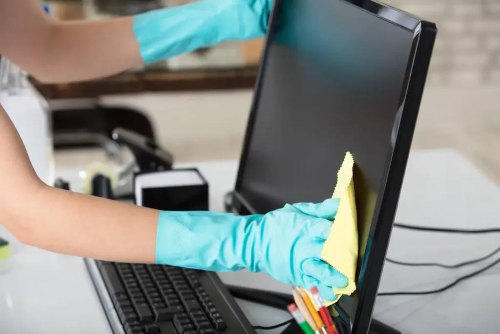 A person wearing teal rubber gloves uses a yellow cloth to clean a computer monitor. A keyboard, smartphone, and colorful pens are visible on the desk.