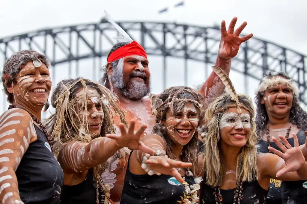 A group of six people wearing traditional attire and body paint stand smiling in front of a large bridge. They have feathers and various accessories in their hair, and one person wears a red headband. They appear joyful, reaching out with open hands.