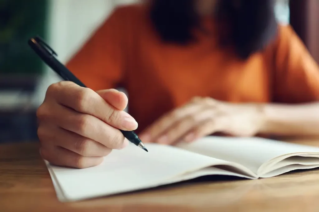 A person in an orange shirt is writing in a notebook with a black pen at a wooden table. The focus is on their hand and the notebook.