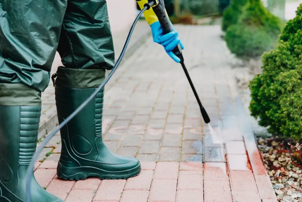 A person wearing green protective clothing, gloves, and boots uses a pressure washer to clean a red brick walkway. Light mist and water spray are visible, with green shrubs lining the path.