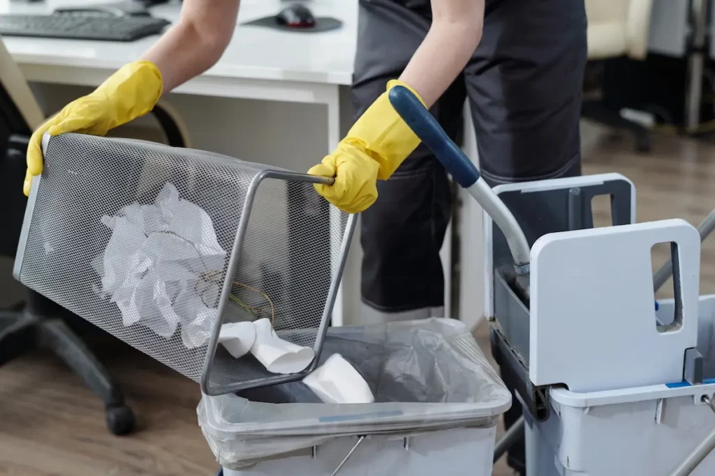 A person wearing yellow gloves is emptying a metal wastebasket filled with crumpled paper into a large trash bin on a cart. The setting appears to be an office with a desk and chair in the background.