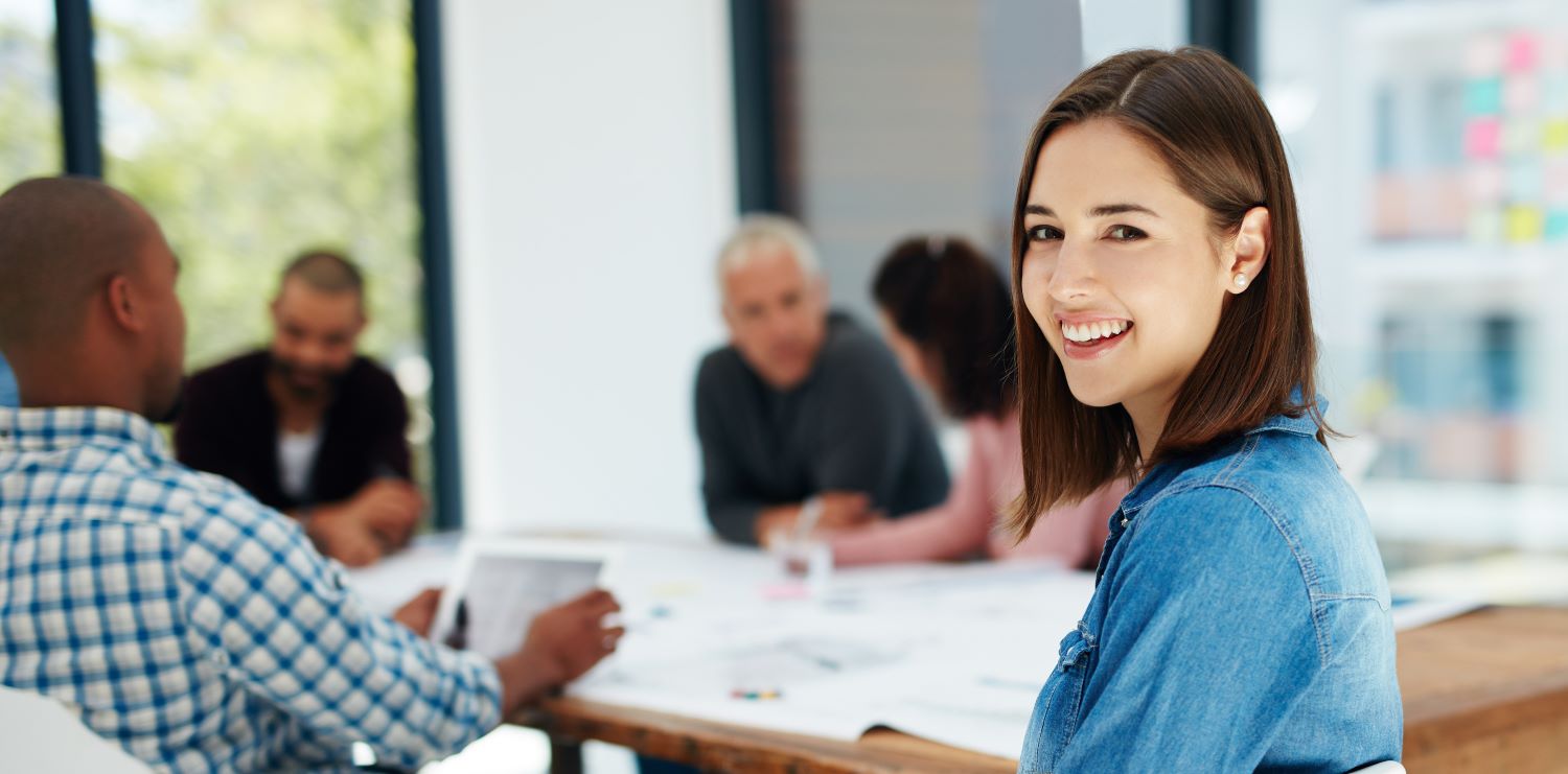 A woman in a denim shirt smiles at the camera while sitting at a table with four other people. The bright, modern conference room reflects workplace cleanliness, with neatly arranged papers on the table. Windows in the background allow natural light to stream in.