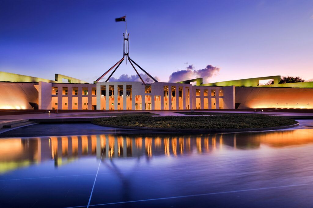 Parliament House in Canberra at dusk glows with warm lights, the symbol of government standing proudly as a flag flutters atop. Its reflection shimmers in the foreground pool under a purple and orange sky, mirroring the clarity brought by innovative cleaning solutions.