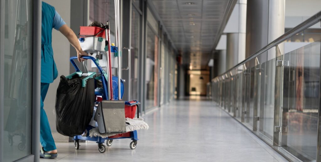 A janitor in blue uniform pushes a cleaning cart down a long, empty hallway with glass railings. The cart is loaded with cleaning supplies, including mops, buckets, and a trash bag. The hallway has a modern design with a polished floor.