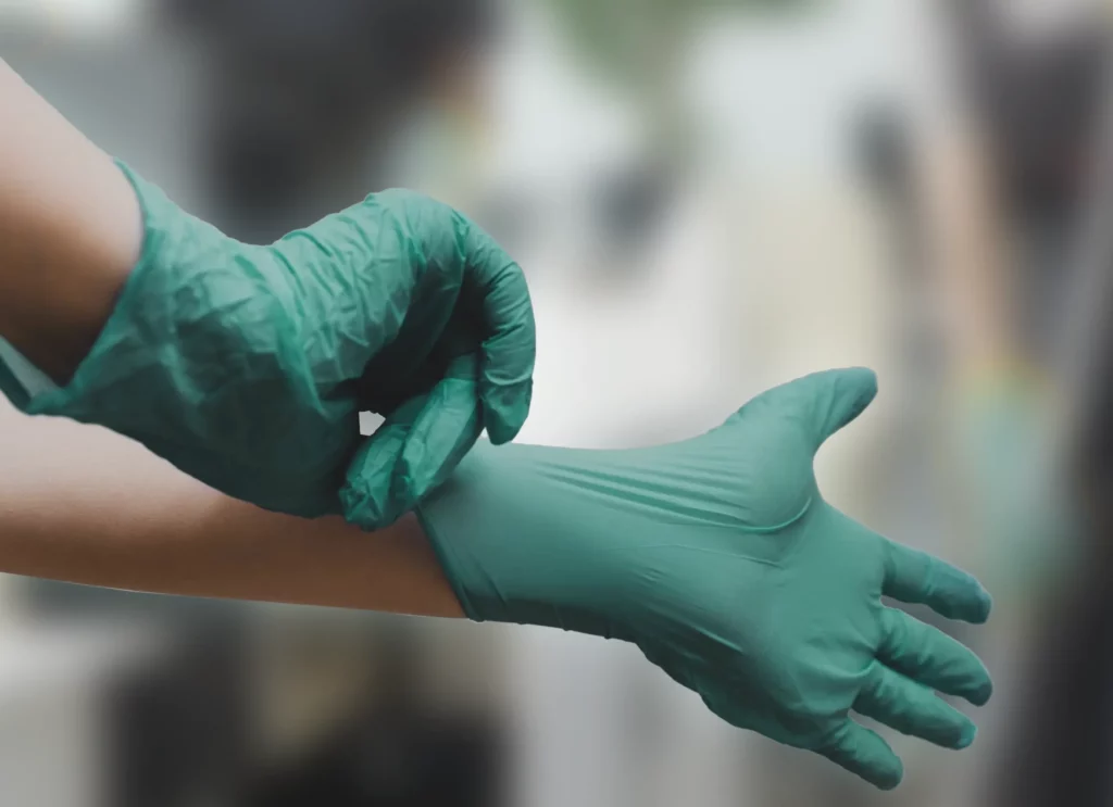 A person is wearing green surgical gloves, pulling one onto their right hand. The background is blurred, highlighting the focus on the hands and gloves.